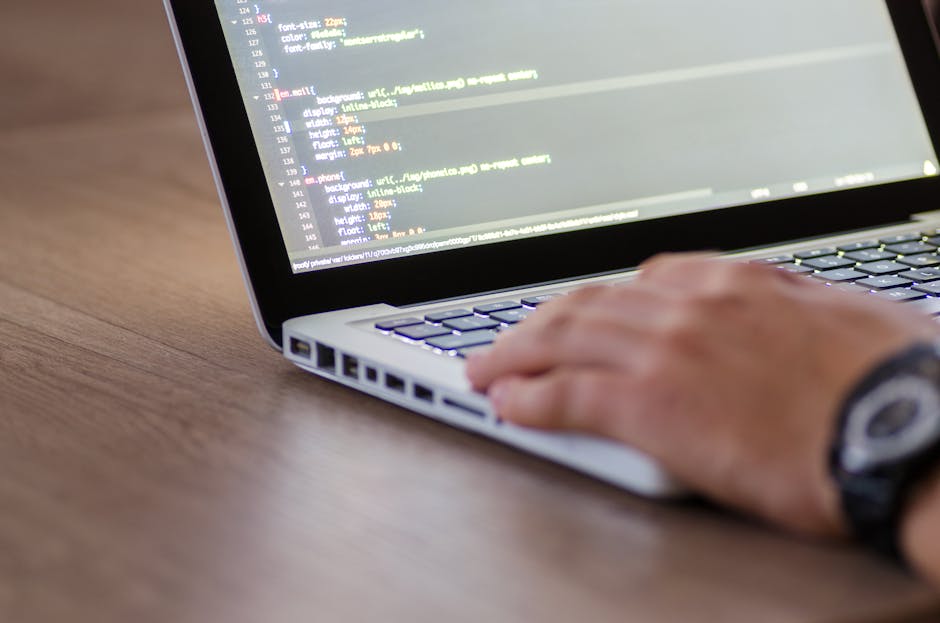 A person's hand with a black wristwatch is positioned on a computer keyboard while coding on the laptop screen, which displays lines of coloured programming code. The laptop, with a silver body and a 