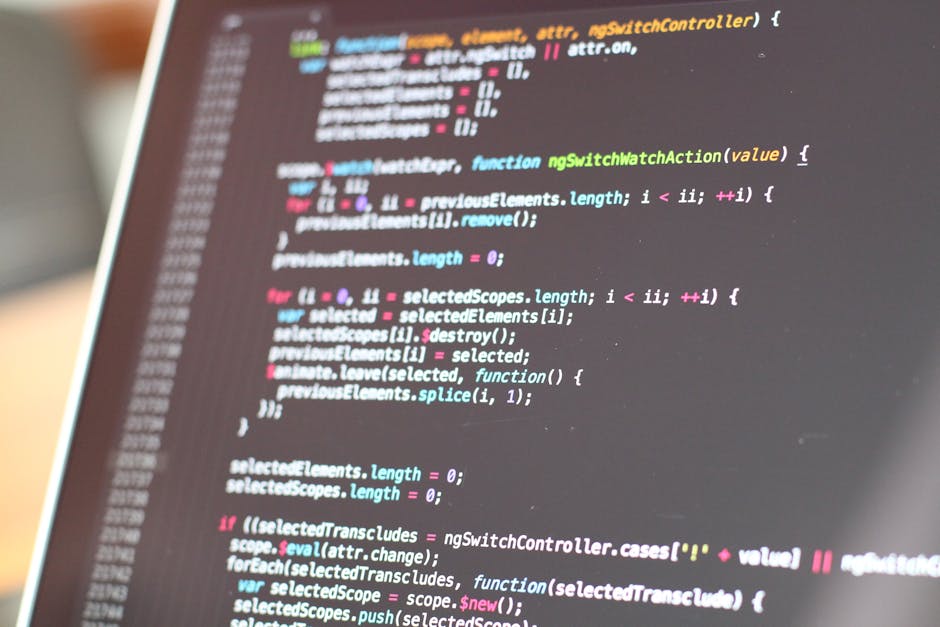 A person's hand with a black wristwatch is positioned on a computer keyboard while coding on the laptop screen, which displays lines of coloured programming code. The laptop, with a silver body and a 